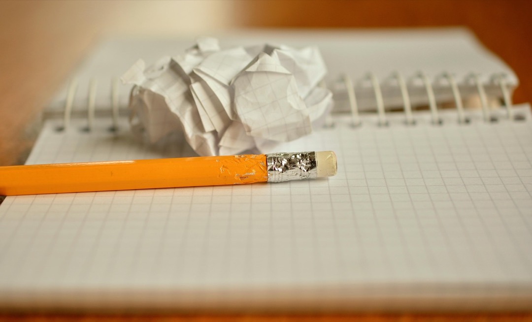 A close up shot of a pencil with a chewed-up ferrule resting on a notebook in front of a crumpled sheet of paper