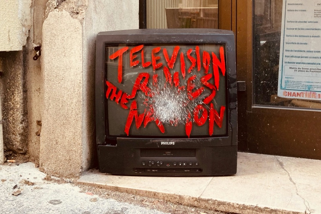 A smashed television sits on the sidewalk in front of a damaged building. The words 'Television rules the nation' have been written on its scratched screen in bright red paint.