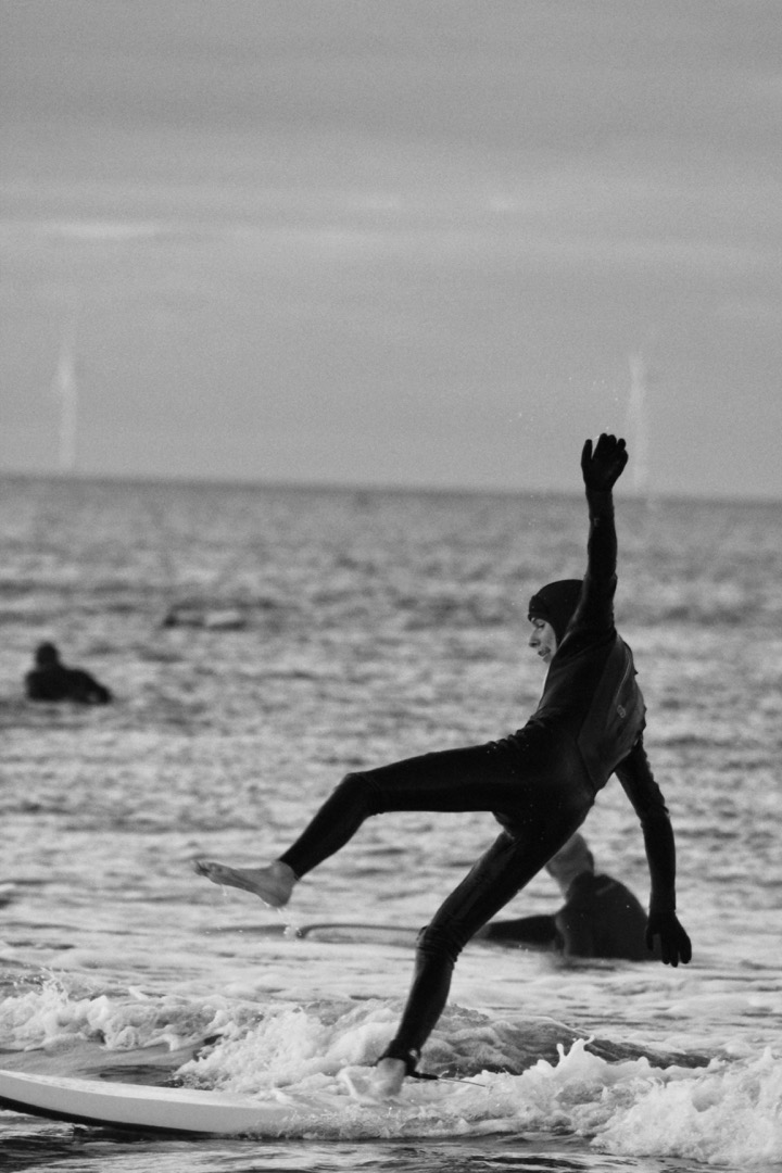 A black and white long-lens shot of a surfer wearing a dark wetsuit frozen in mid-fall from their board with arms splayed and one foot in the air