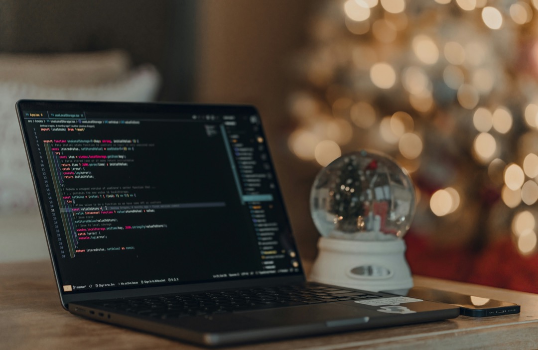 A laptop whose screen displays code in an editor sitting on a desk with a phone and a snowglobe, with a lit Christmas tree in the background