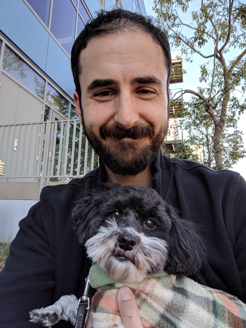 A picture of me looking more or less into the camera holding a black and white Havanese wearing a little plaid sweater